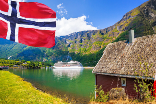 Links eine norwegische Fahne, im Hintergrund im einem Fjord gelegen ein wei&szlig;es Kreuzfahrtschiff, rechts ein rotes Holzhaus